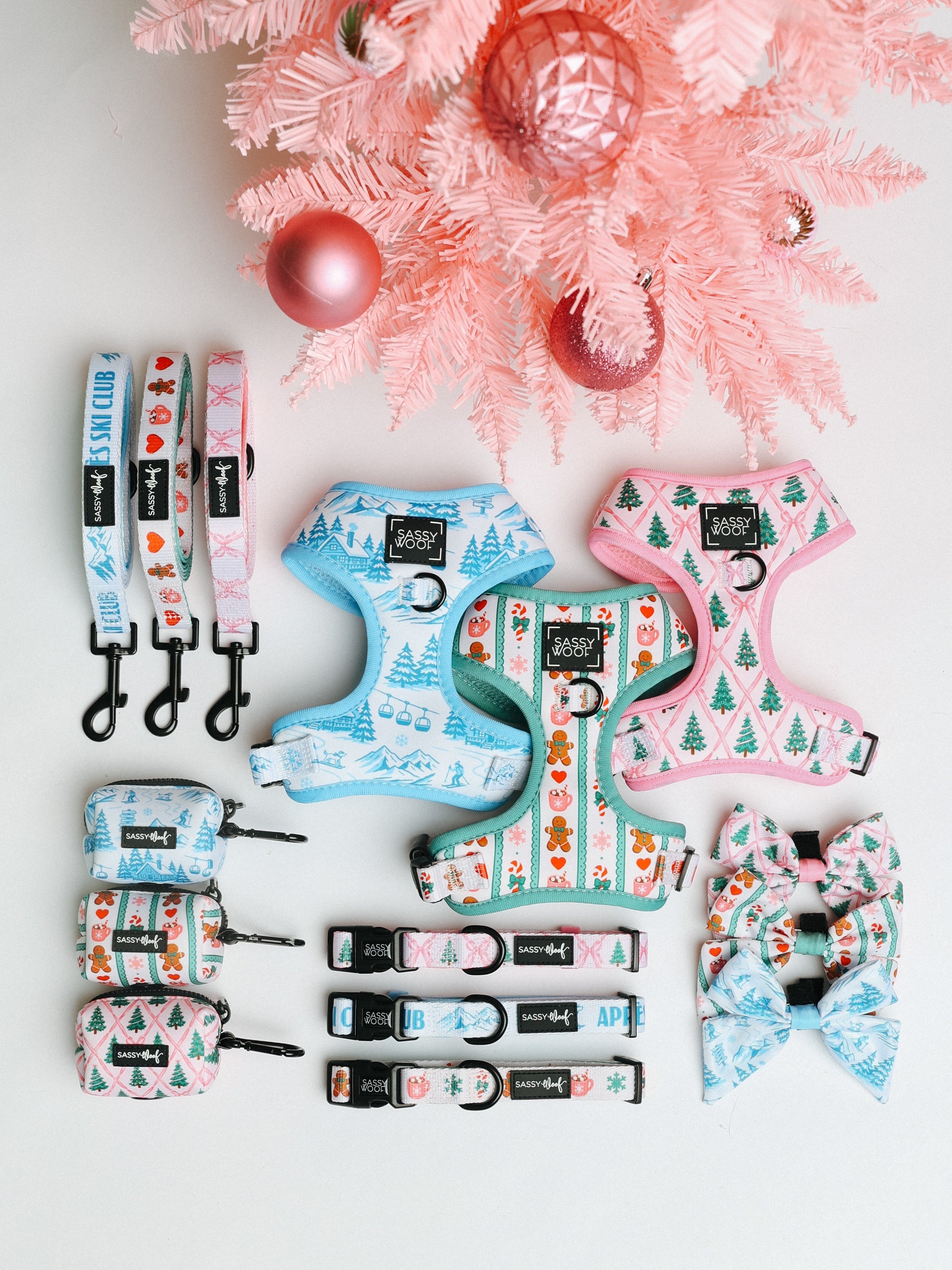 Dog accessories, including leashes, harnesses, collars, bows, and bag holders, with pastel holiday patterns are arranged under a pink Christmas tree with pink ornaments.