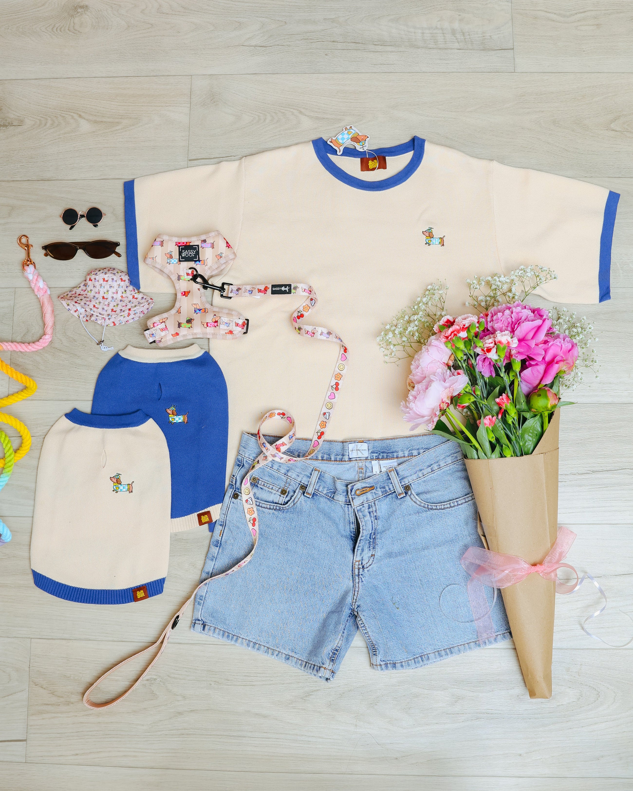 Clothes, accessories, and flowers on a wooden floor: a cream T-shirt with blue trim, matching dog shirts, denim shorts, sunglasses, a leash, scrunchies, and a bouquet of pink flowers.