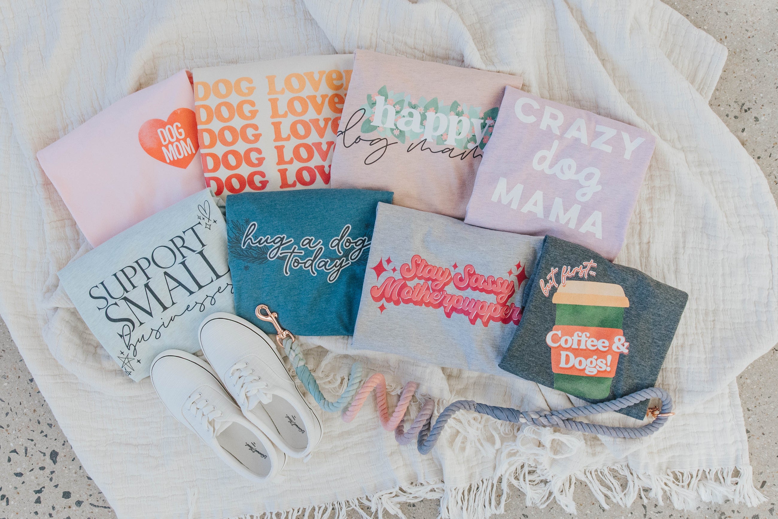 A collection of colorful dog-themed t-shirts, a pair of white sneakers, and two leashes arranged on a white textured blanket on a speckled floor.