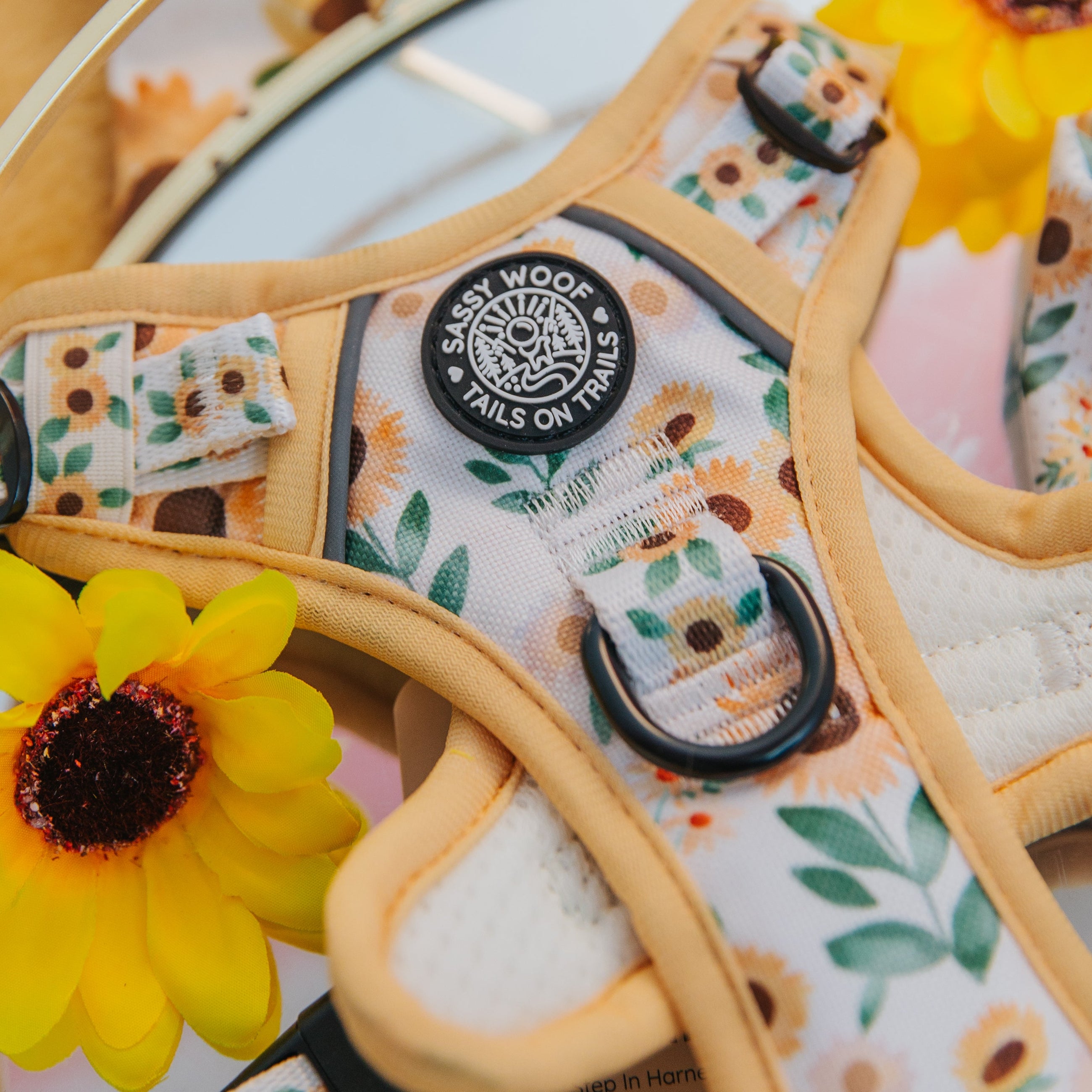 Close-up of a floral-patterned dog harness with Sassy Woof Tails on Trails on a black tag, surrounded by bright yellow artificial sunflowers.