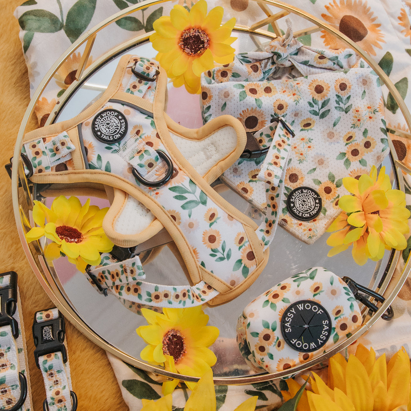 A flat lay of dog accessories—harness, leash, waste bag holder, and bandana—all featuring a sunflower pattern, surrounded by fake sunflowers and floral fabric on a round tray.