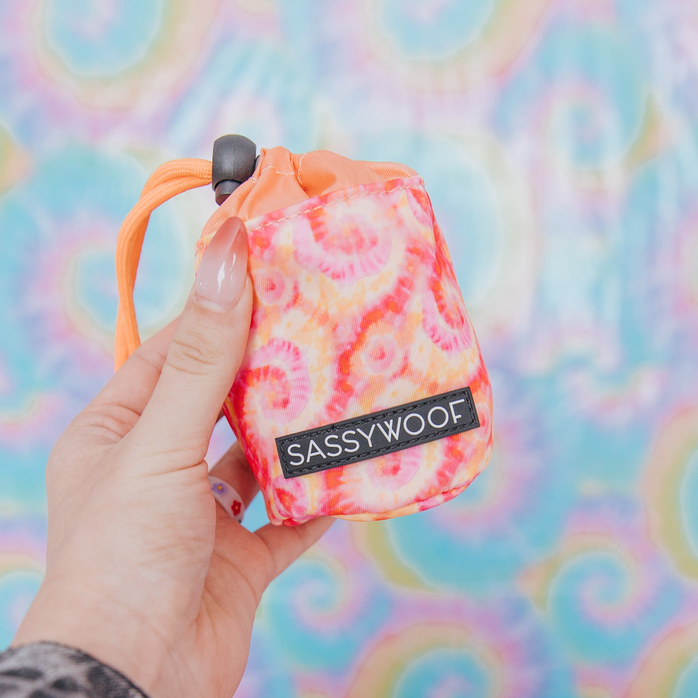 A hand with painted nails holds a colorful tie-dye pouch labeled SASSYWOOF against a bright, rainbow tie-dye background.