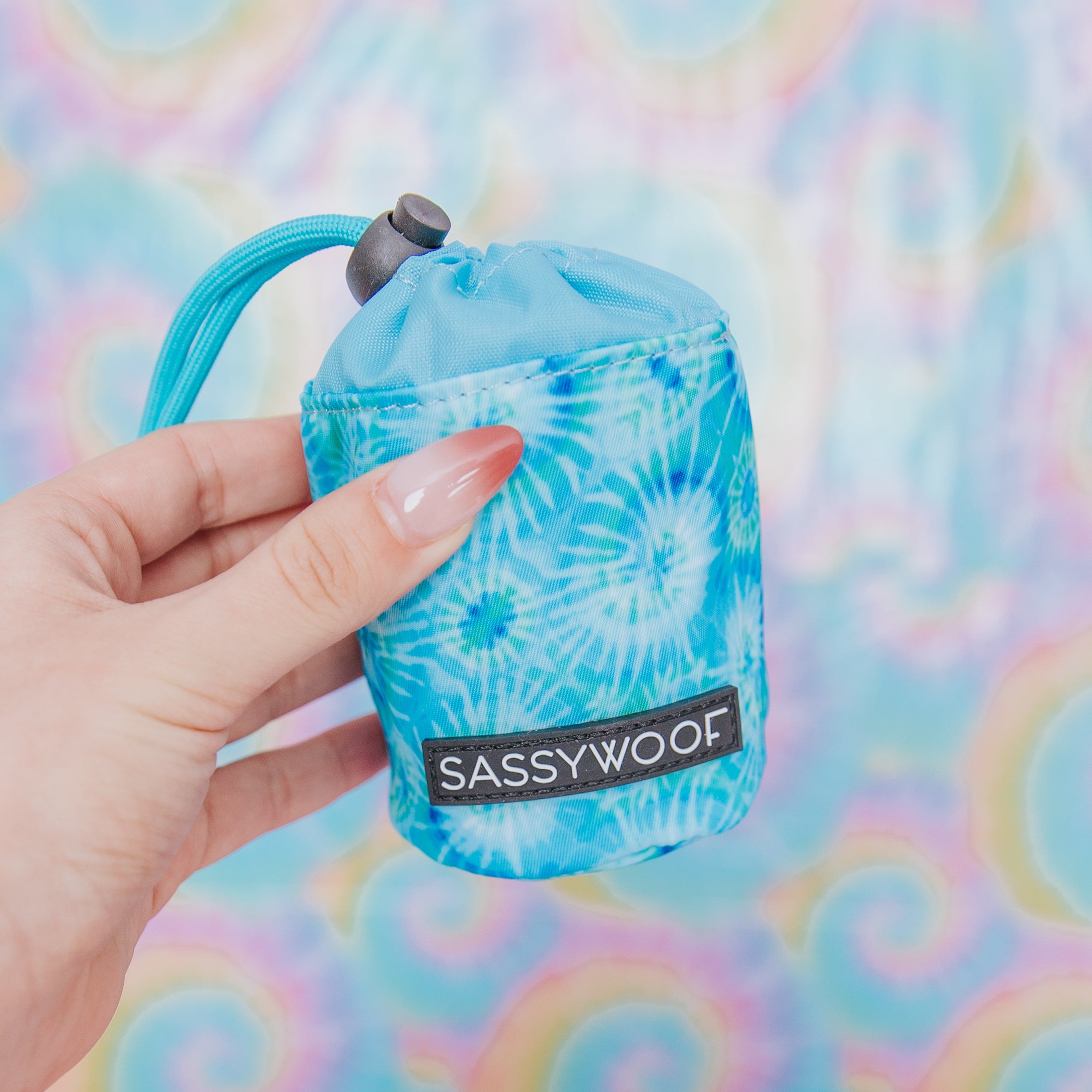 A hand with long, manicured nails holds a small blue tie-dye pouch labeled SASSY WOOF against a colorful, swirled pastel background.