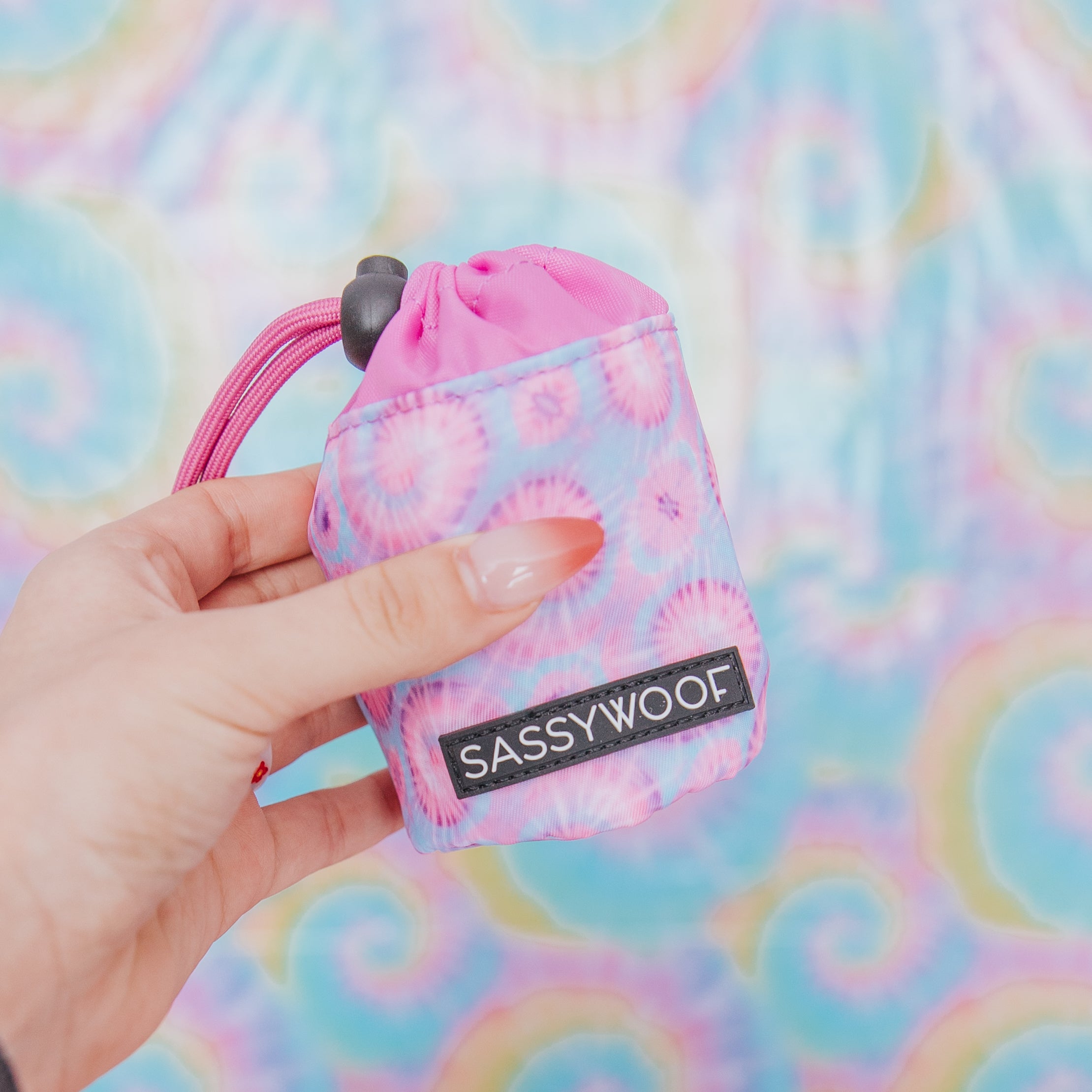 A hand holds a colorful tie-dye pouch with a SASSYWOOF label against a matching pastel tie-dye background.