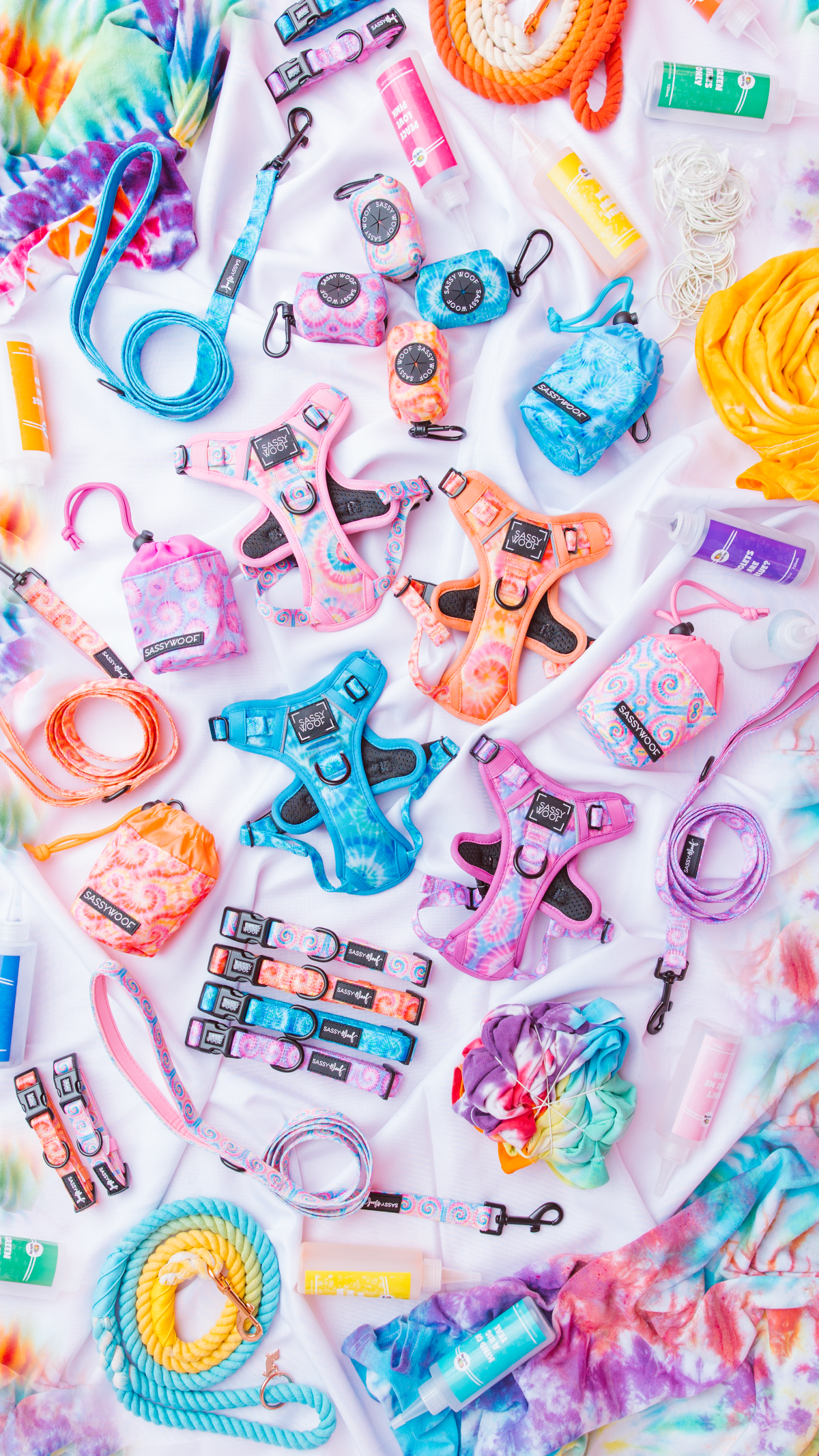 Assorted colorful dog harnesses, leashes, collars, waste bag holders, and grooming products arranged on a vibrant, tie-dye fabric background.