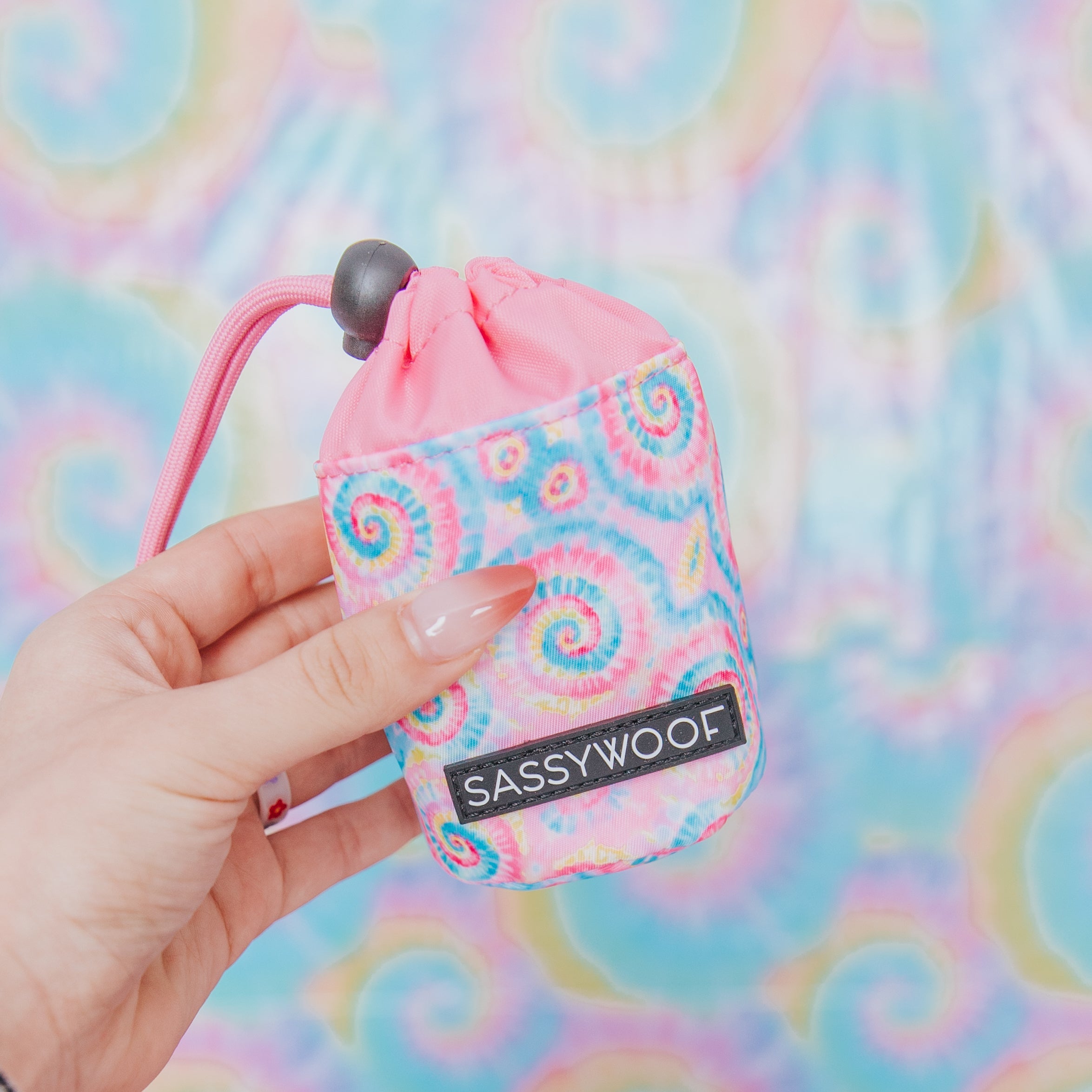 A hand holding a colorful tie-dye pouch labeled “SASSY WOOF” with a pink drawstring, against a matching tie-dye background.