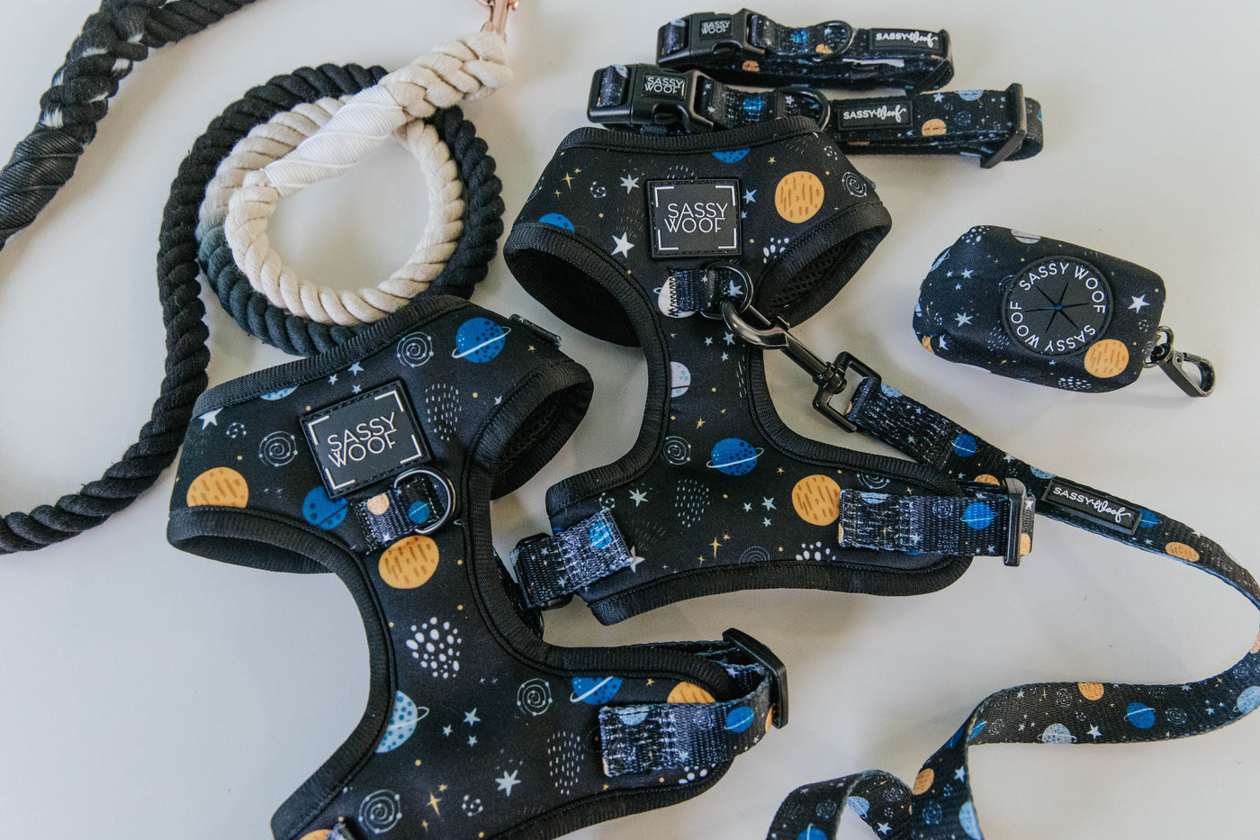 A set of dog accessories with a space-themed design, including harnesses, leashes, collars, and a waste bag holder, all decorated with planets and stars on a black background.