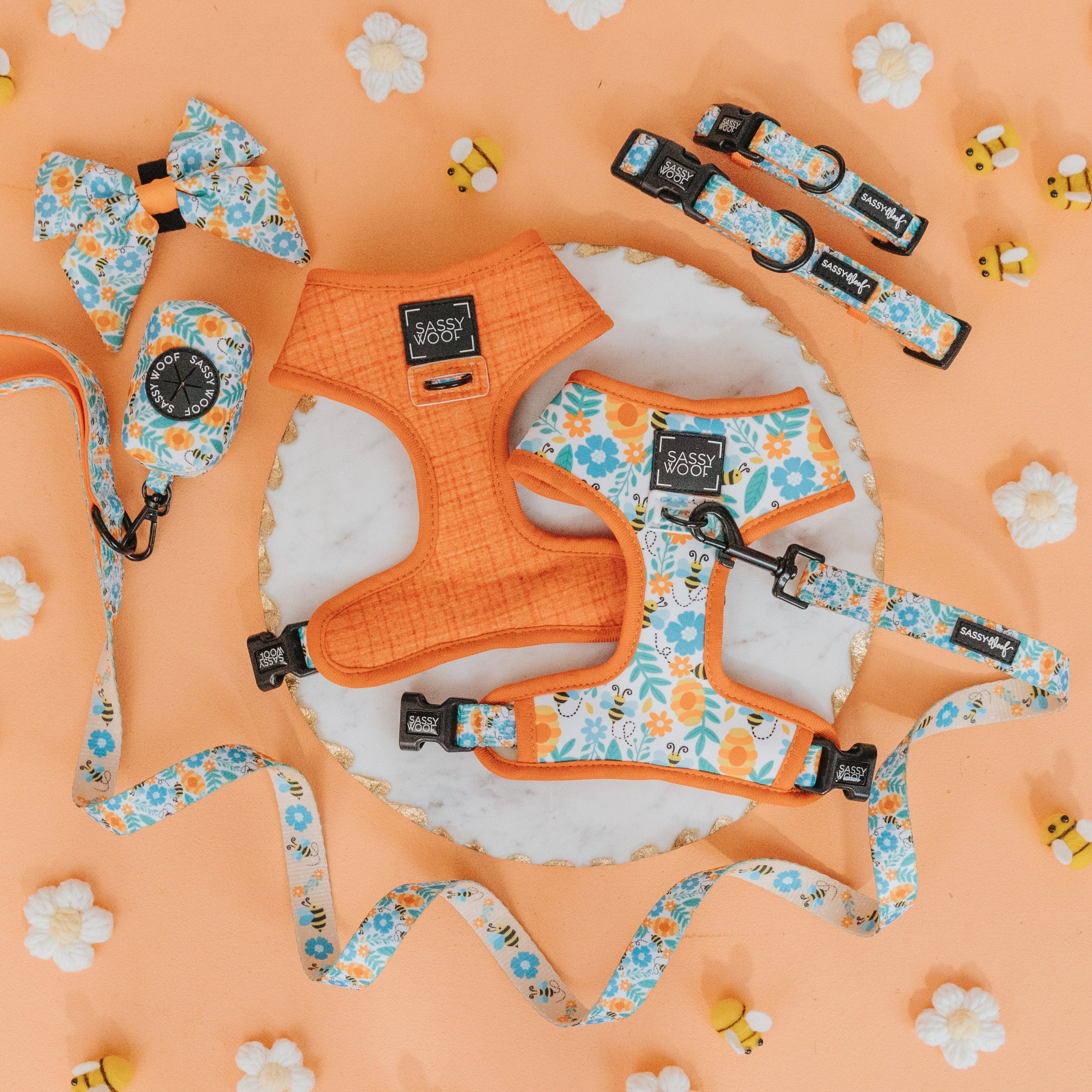 Floral and orange dog harnesses, leashes, collars, bow ties, and waste bag holders are arranged on a peach background with decorative flowers and bees.