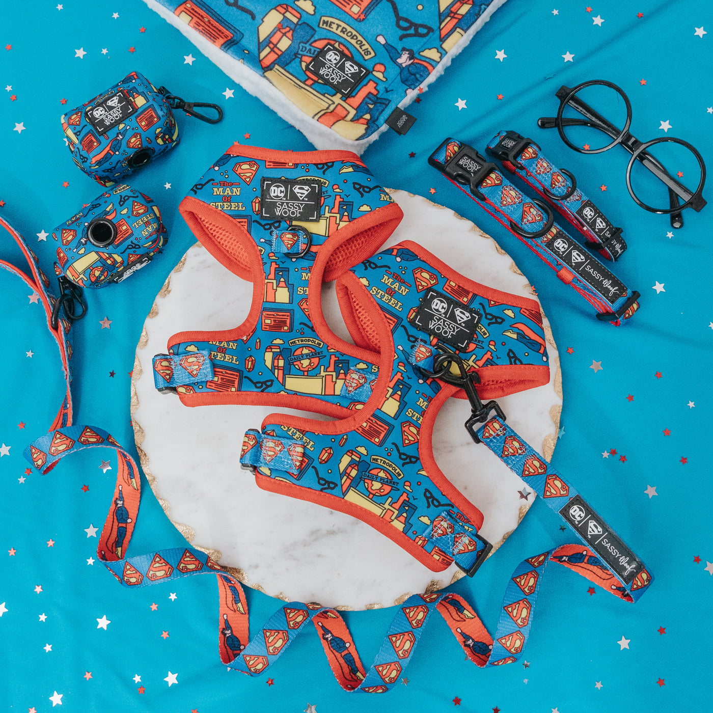 A collection of dog accessories with a blue and red Superman comic print, including a harness, leash, collar, poop bag holder, and a dog tag, arranged on a blue star-patterned background.