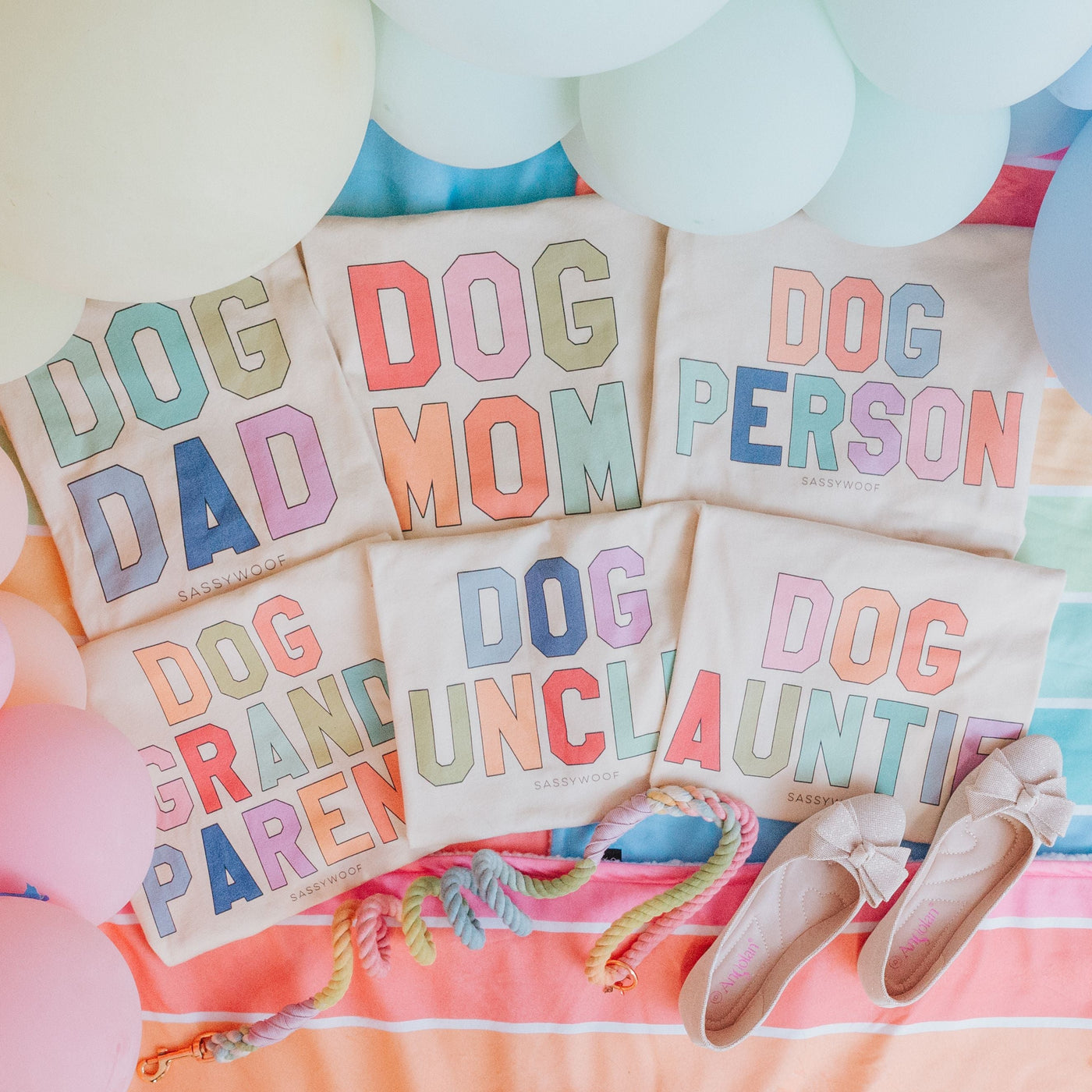 Five pastel-colored t-shirts with bold text: Dog Dad, Dog Mom, Dog Person, Dog Grandparent, Dog Uncle, and Dog Auntie are laid out on a rainbow towel with balloons and shoes.