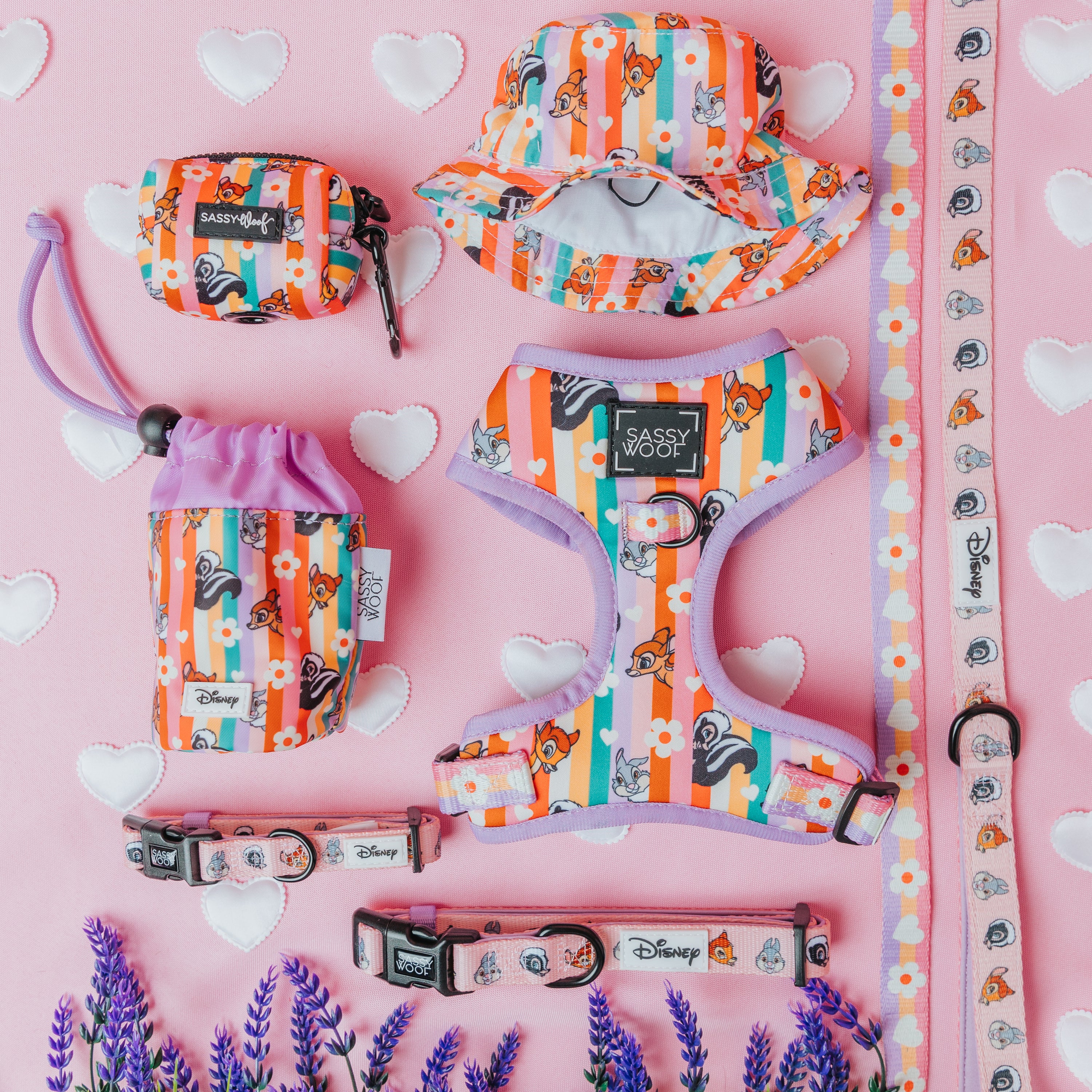 Colorful Disney-themed pet accessories, including collars, harness, leash, poop bag holders, and a bucket hat, are arranged on a pink background with heart accents and lavender sprigs.