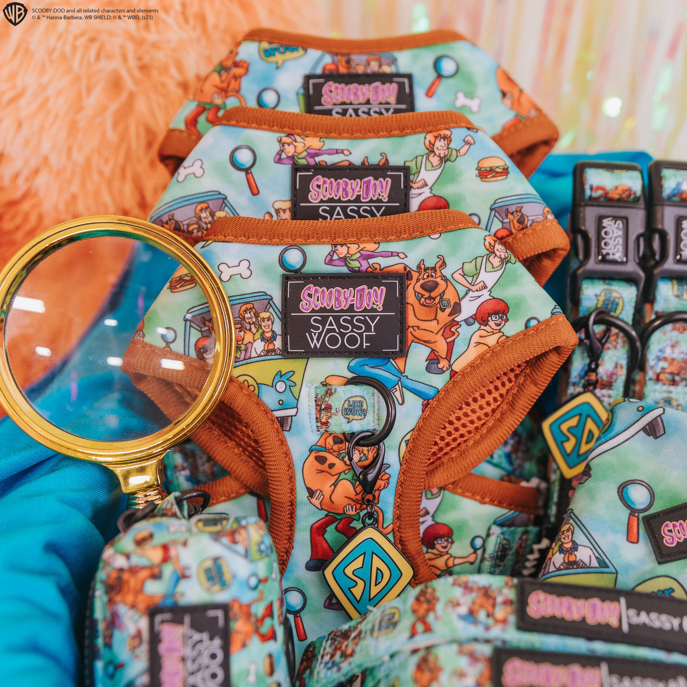 Colorful dog harnesses, leashes, and accessories featuring Scooby-Doo cartoon characters are neatly displayed with a magnifying glass on top, against a bright, fuzzy orange pillow background.