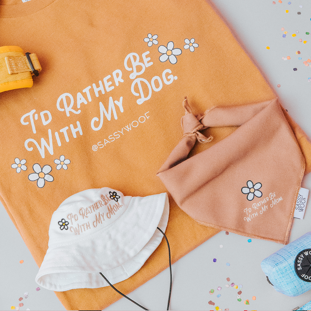 Flat lay of Sassy Woof’s Human Tee - I’d Rather Be With My Dog (printed with eco-friendly ink), dog mom bucket hat, blue & orange bandanas, and a floral orange dog harness on a light surface.