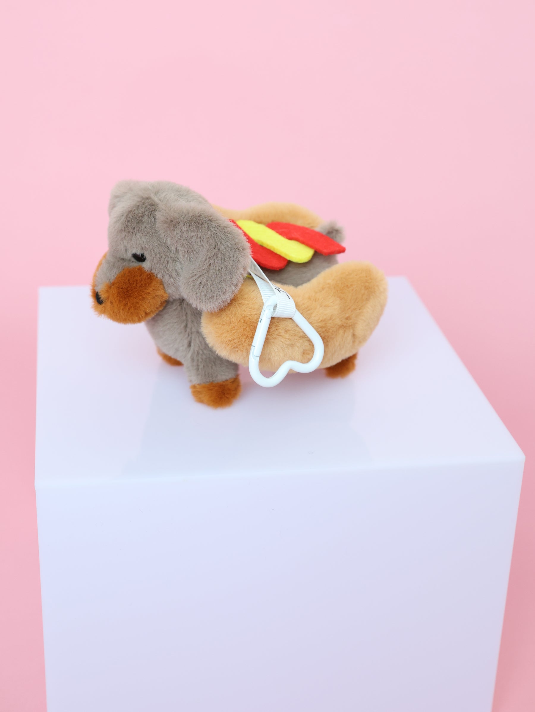 A Sassy Woof Blind Box collectible dachshund plush keychain, dressed as a hot dog with bun, mustard, and ketchup, stands on a white cube against a pink background. A white heart-shaped clip is attached to its side.