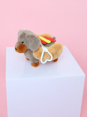 A Sassy Woof Blind Box collectible dachshund plush keychain, dressed as a hot dog with bun, mustard, and ketchup, stands on a white cube against a pink background. A white heart-shaped clip is attached to its side.