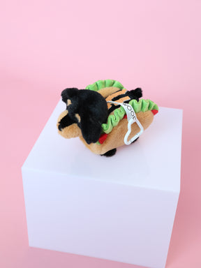 A Sassy Woof Blind Box plush keychain shaped like a dachshund in a hot dog costume with green lettuce-like fabric, shown on a white cube against a pink background.