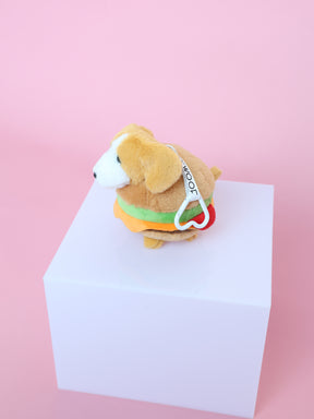 A Sassy Woof Blind Box collectible plush, styled as a dog in a hamburger costume with a yellow bun, green lettuce, red tomato details, and a “woof” tag, sits on a white cube against a pink background.