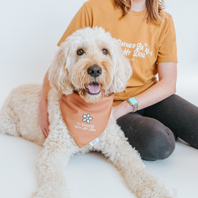 A fluffy white dog in a tan bandana sits next to a person wearing the Human Tee - I’d Rather Be With My Dog by Sassy Woof, both featuring a small flower and eco-friendly ink. The person’s hand rests on the dog’s back.
