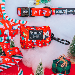 Sassy Woof’s Dog Waste Bag Holder - Peanuts™ Beagle Bells, featuring Snoopy and festive Christmas designs, is displayed on snow-like fabric with mini pine trees and a wrapped gift.