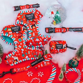 Sassy Woof’s Dog Four Piece Bundle - Peanuts™ Beagle Bells, featuring red, green, and white holiday patterns, is displayed among fake snow, mini Christmas trees, ribbon, candy canes, and a snow globe.
