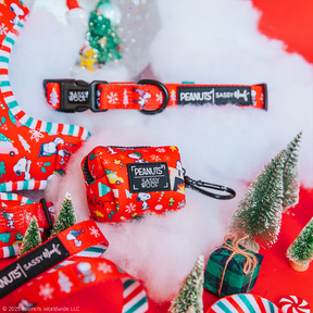 The Sassy Woof Collar Three Piece Bundle - Peanuts™ Beagle Bells is displayed on white fluffy material among mini Christmas trees, gifts, and candy canes for a festive touch.