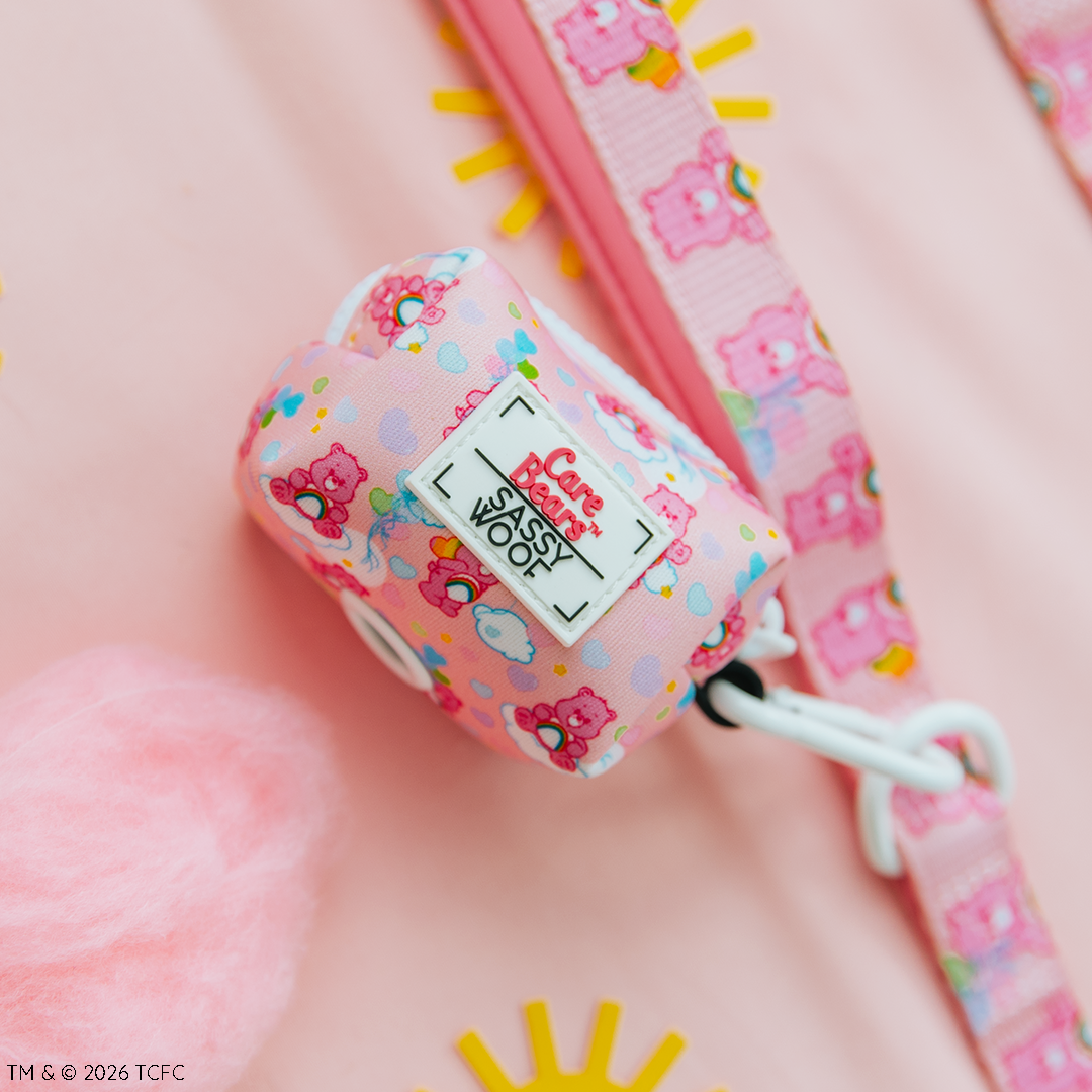The Sassy Woof Dog Waste Bag Holder – Care Bears™ Cheer Bear, featuring pink cartoon bear prints, is displayed on a matching pink harness and leash amid sun shapes and fluffy decor on a pastel background.