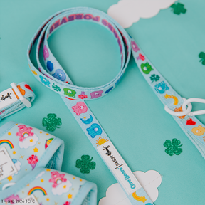 The Sassy Woof Collar Three Piece Bundle - Care Bears™ Care-a-Lot features nostalgic pet accessories with playful character faces and rainbows, displayed on a teal surface with white clouds and green glittery shamrocks.