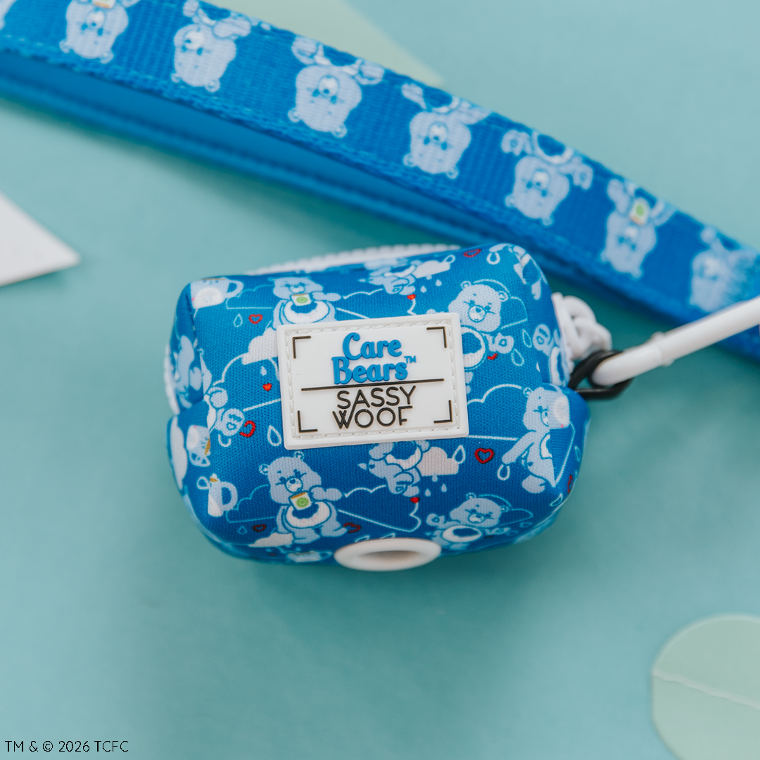 The Sassy Woof Dog Waste Bag Holder - Care Bears™ Grumpy Bear, shown with a matching leash and pet harness featuring blue and white Grumpy Bear designs, is displayed on a light blue background.