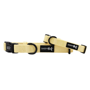 The Sassy Woof Dog Collar - Daffodil is a pale yellow, adjustable collar with black plastic buckles, two black metal rings, and “SASSY WOOF” labels. The collar is shown laid flat with one end slightly overlapping the other.