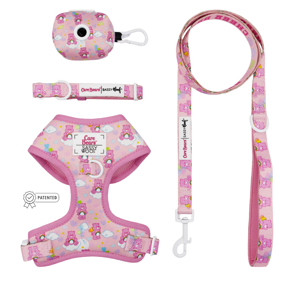 The Sassy Woof Dog Four Piece Bundle - Care Bears™ Cheer Bear includes a pink adjustable harness, leash, and waste bag holder with white clasps and metal clips, featuring colorful Care Bears details—ensuring comfort as your puppy grows!.