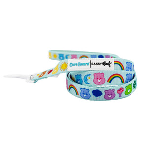 The Sassy Woof Collar Three Piece Bundle - Care Bears™ Care-a-Lot features a light blue design with colorful Care Bears faces, rainbows, suns, and moons, plus “Care Bears” and “Sassy Woof” logo tags near the white clasp.