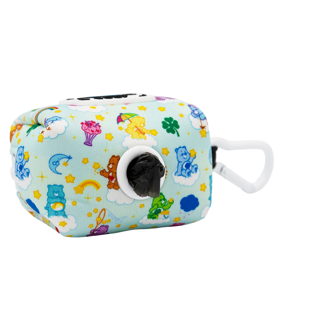 The Sassy Woof Dog Waste Bag Holder - Care Bears™ Care-a-Lot features a rectangular design with clouds, stars, rainbows, and Care Bears. It has a black bag in the opening and a white plastic clip for easy attachment.
