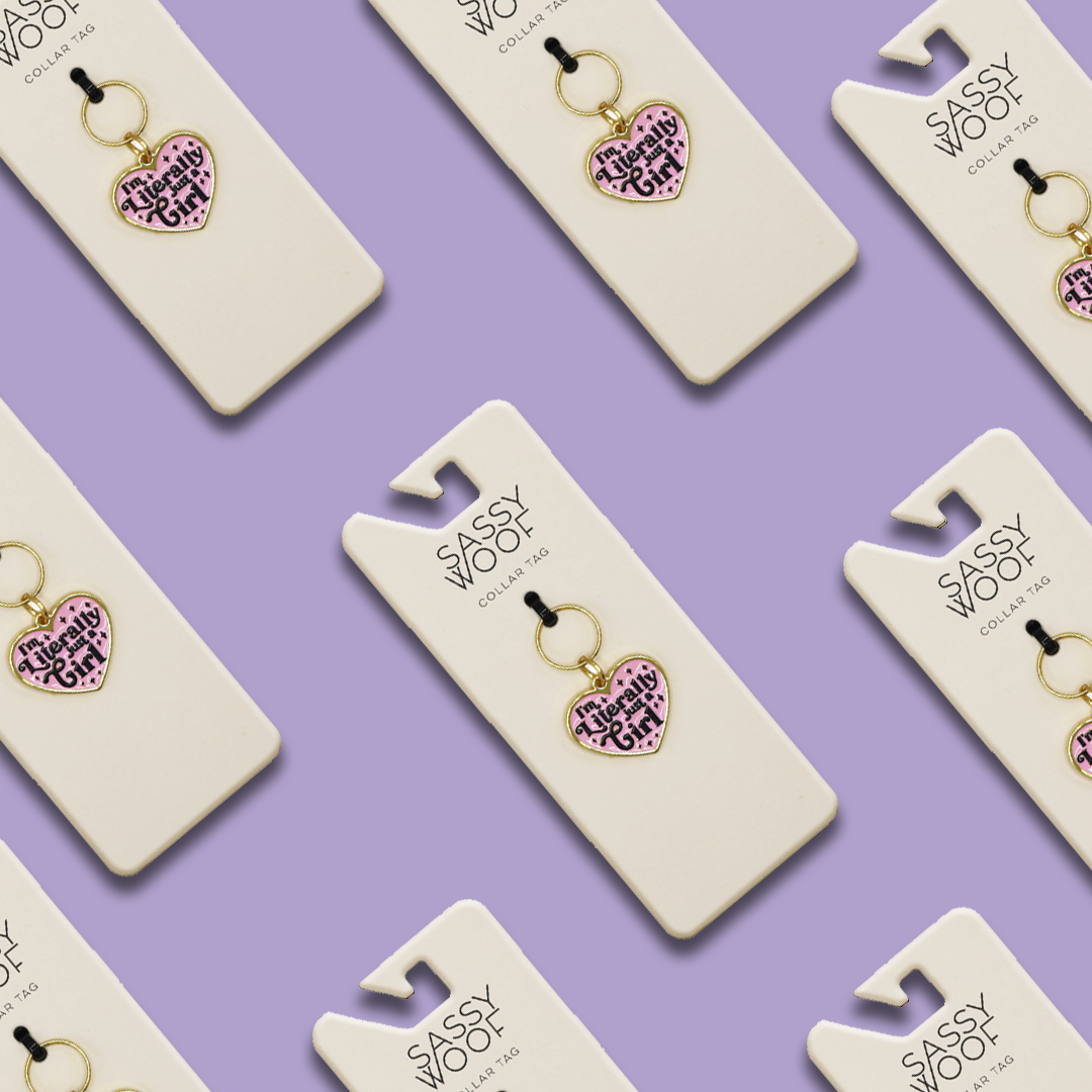 Sassy Woof Collar Tag - Just a Girl, featuring pink heart shapes and gold accents with the phrase "This is my selfie side," is displayed diagonally on branded cards against a light purple background.