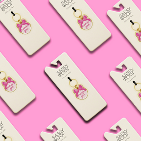 Flat lay of multiple Sassy Woof Collar Tag - Mama's Girl gold keychains with pink bows, displayed on white cards against a pink background.
