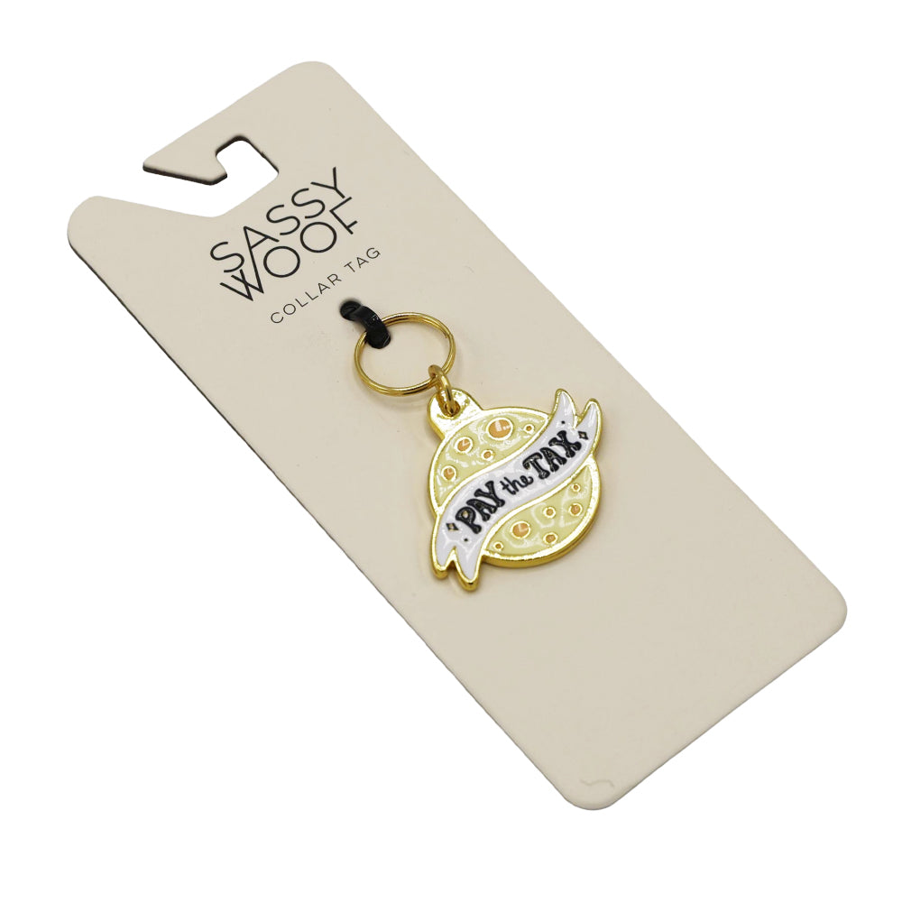 The Sassy Woof Collar Tag - Cheese Tax is a gold and white enamel dog charm shaped like a planet with “pay the tax” text, attached to a branded cream paper card—perfect for your Cheese Tax Collector.