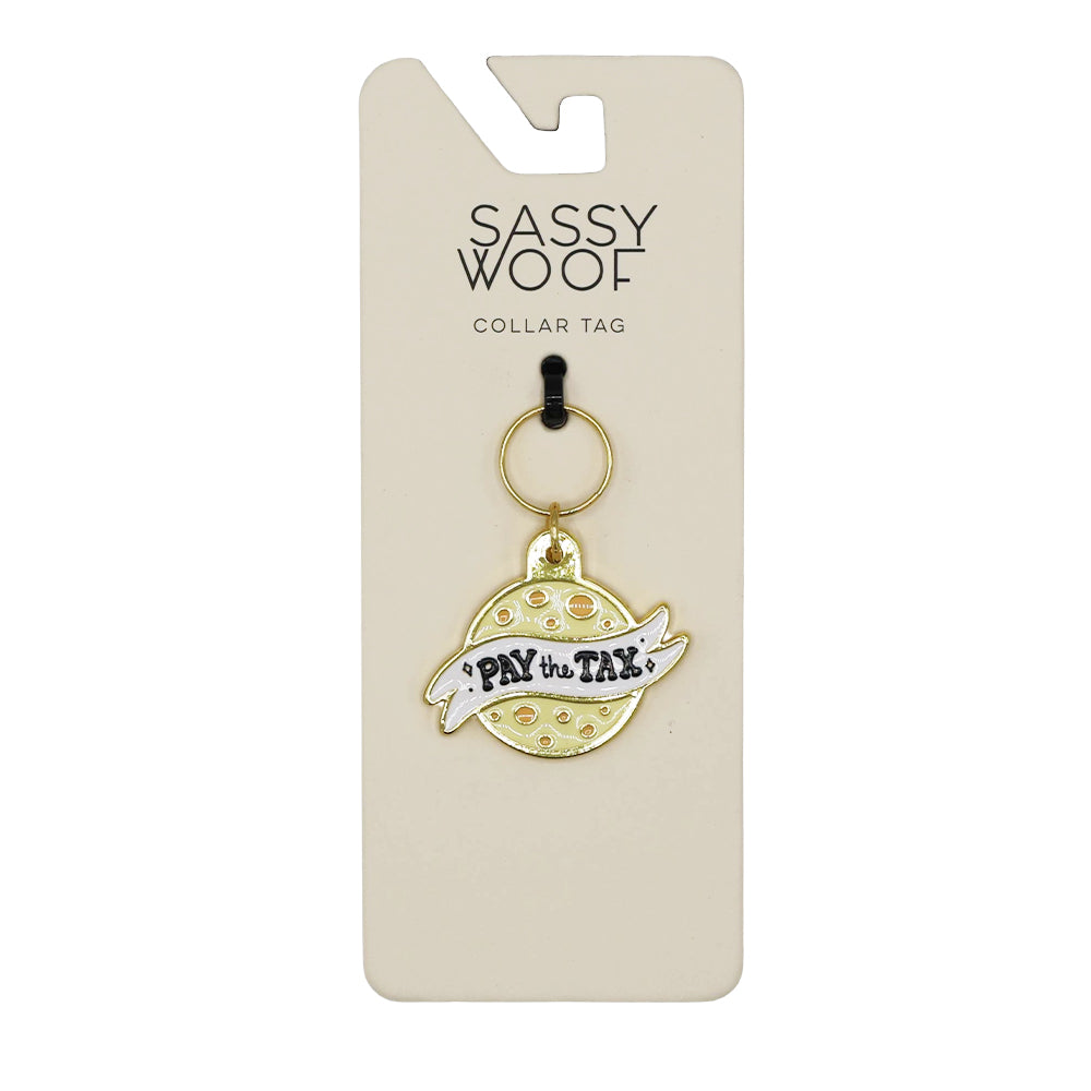 The Sassy Woof Collar Tag - Cheese Tax is a playful gold and white dog charm shaped like a planet with a ring, featuring "PAY the TAX," attached to a card labeled SASSY WOOF COLLAR TAG.