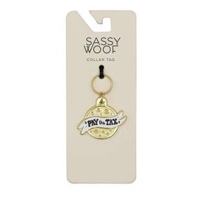 The Sassy Woof Collar Tag - Cheese Tax is a playful gold and white dog charm shaped like a planet with a ring, featuring "PAY the TAX," attached to a card labeled SASSY WOOF COLLAR TAG.