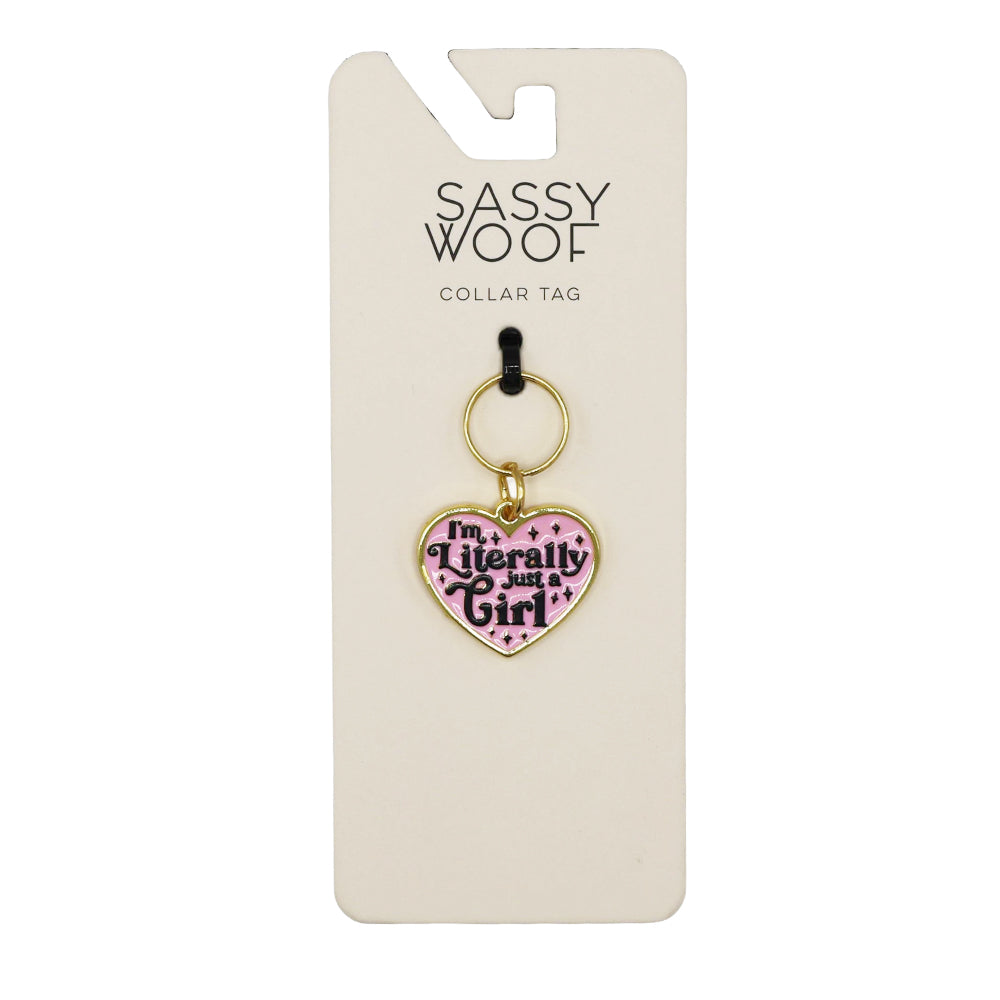 The Sassy Woof Collar Tag - Just a Girl is a pink, heart-shaped dog tag with gold accents and "Im Literally Just a Girl" in black, attached to a keyring and displayed on branded packaging.