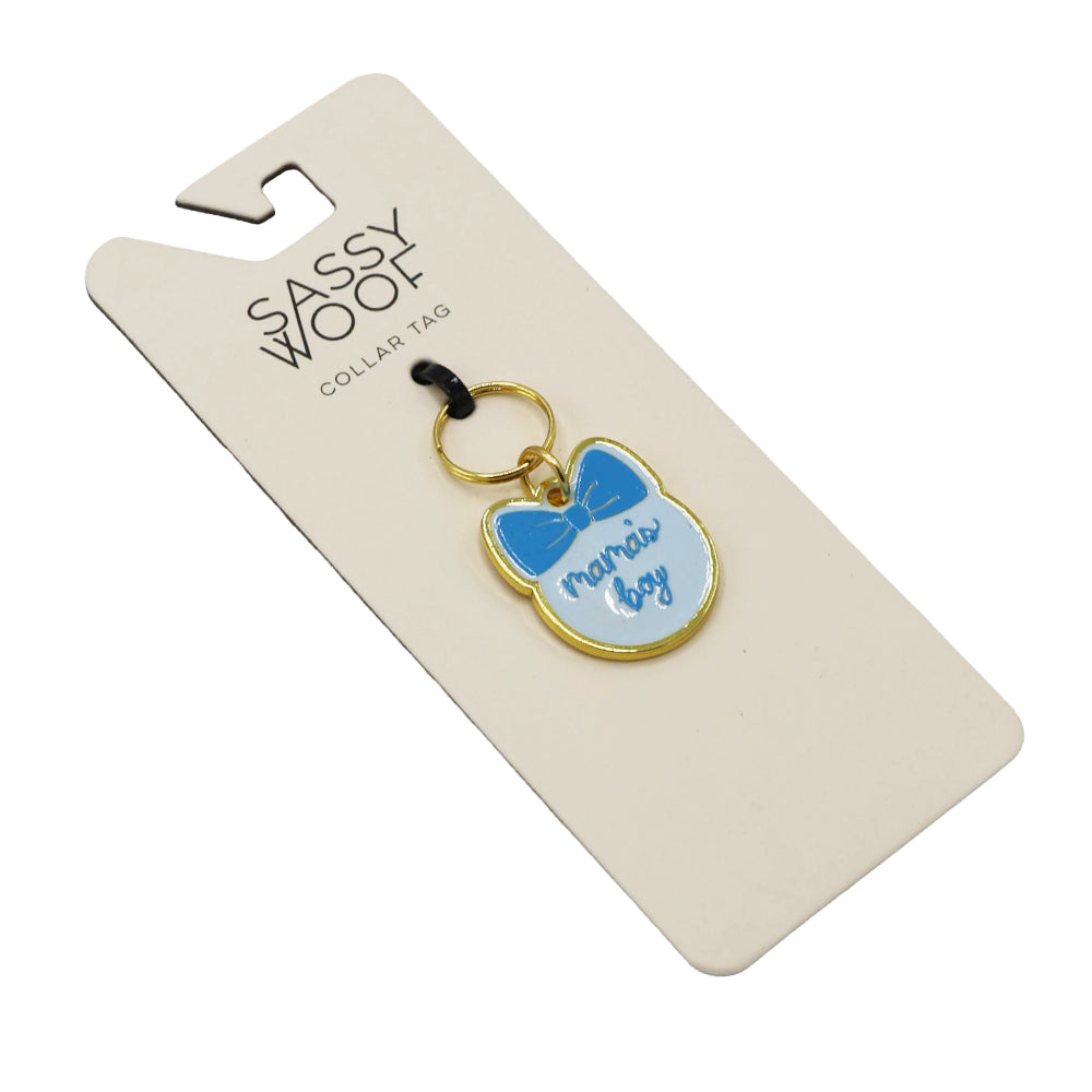The Sassy Woof Collar Tag - Mama's Boy is a gold dog collar charm with a blue bow and “mamas boy” in blue text, attached to a branded card with a black ring—perfect for any proud dog mom.
