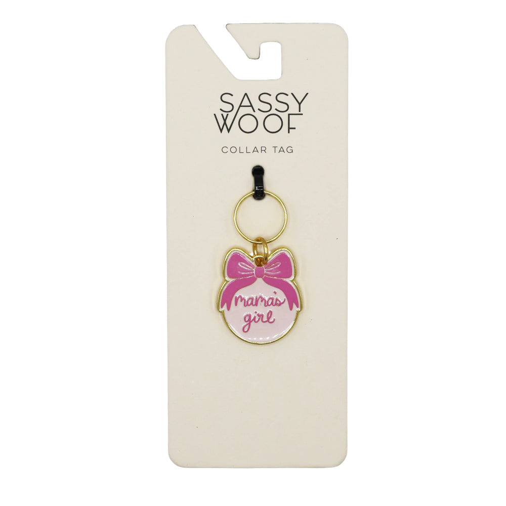 The Sassy Woof Collar Tag - Mama's Girl features a round design with a pink bow and “mamas girl” in pink script, hanging from a gold keyring to add charm to any pup’s look.
