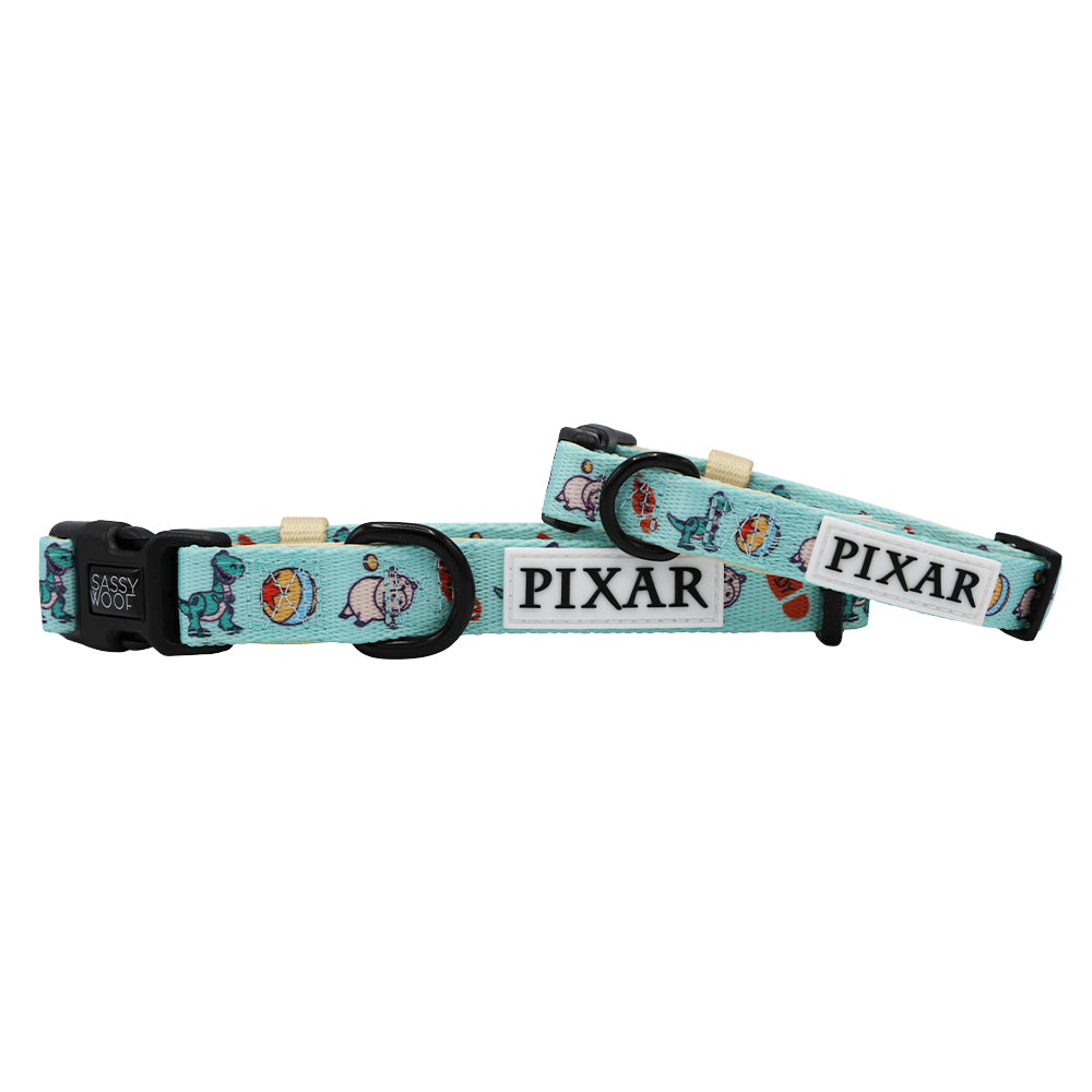 Two light blue Sassy Woof dog collars, adjustable with black buckles, feature Disney and Pixar's Toy Story characters and icons including a dog, duck, and Pixar logo on white patches. These collars are stacked together.