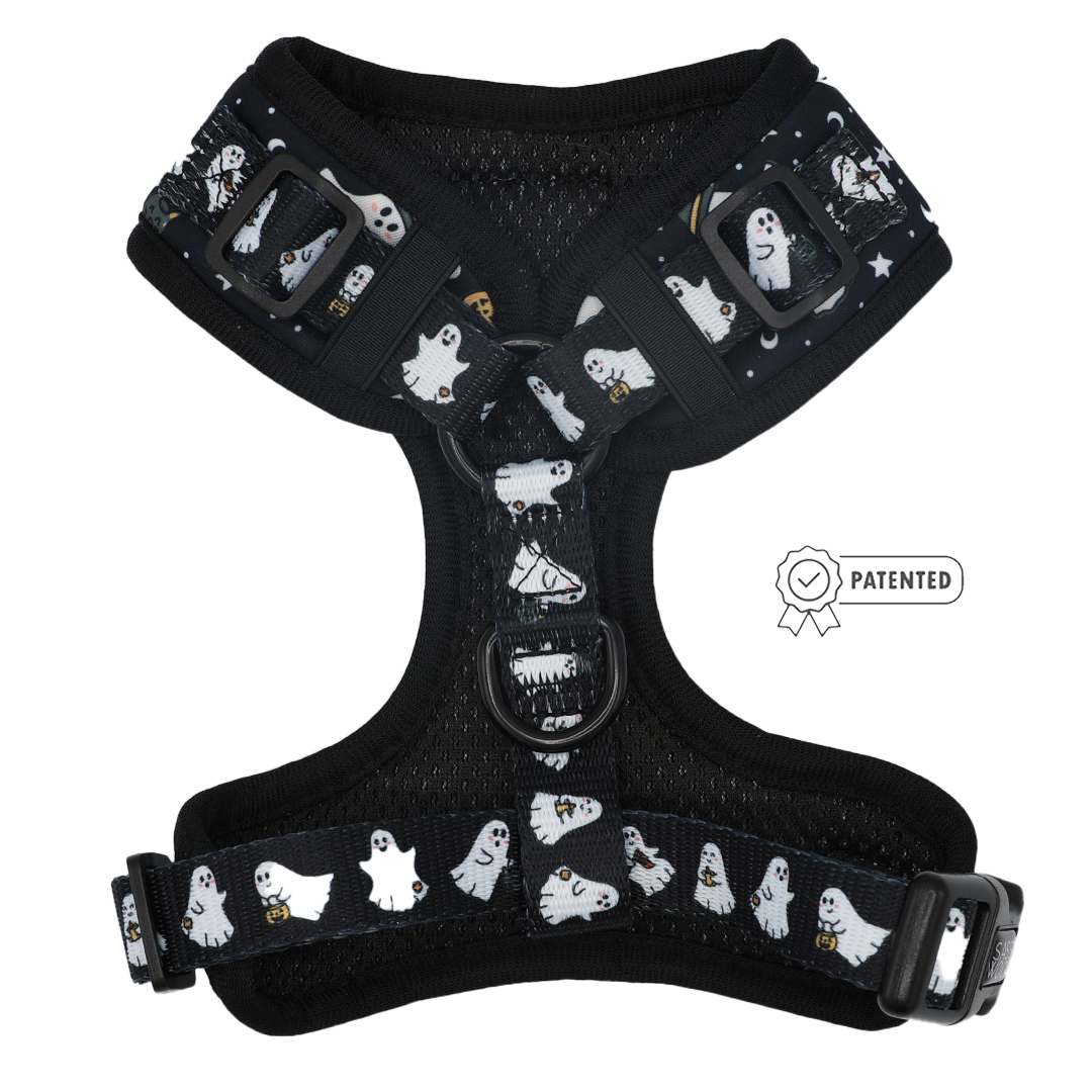 The Sassy Woof Dog Adjustable Harness - Lil Boo Thing is a black harness adorned with white cartoon ghosts, some holding lanterns, and printed stars. This Halloween dog harness features adjustable straps, sturdy metal rings, durable plastic buckles, and a Patented badge icon on the right side.