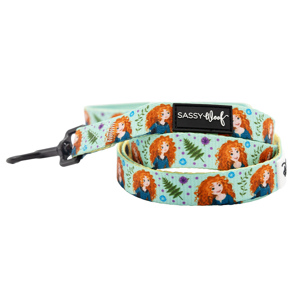 The Sassy Woof Harness Three Piece Bundle – Disney's Merida features a dog leash with a light blue background, cartoon Merida in a blue dress, green leaves, purple flowers, and a black Sassy Woof label.