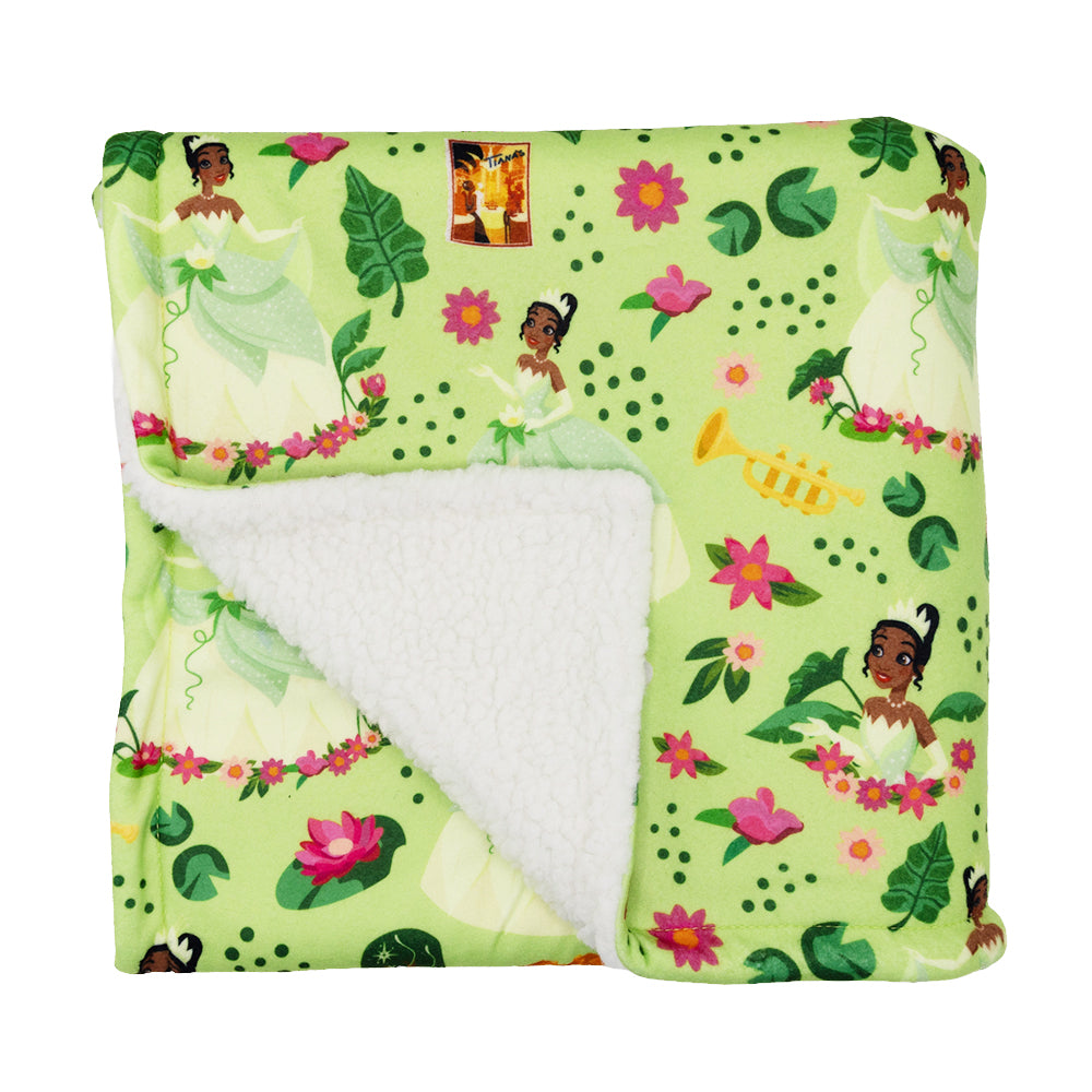 The Sassy Woof Pet Blanket - Disney's Tiana is a folded green fleece sherpa dog blanket with white sherpa lining, featuring Princess Tiana, pink flowers, green leaves, lily pads, and a yellow trumpet.