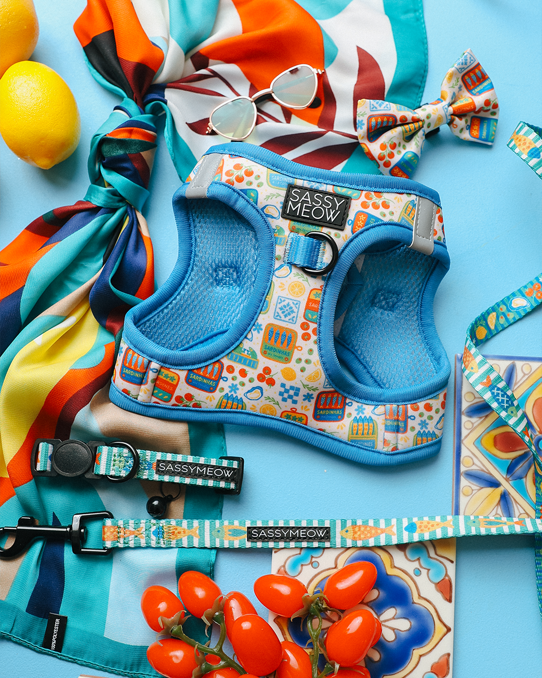 A vibrant flat lay shows the Sassy Woof Cat Bundle - Portofino: a blue harness, leash, and collar with colorful patterns, plus a matching bow tie, sunglasses, multicolored scarf, lemons, tomatoes, and tile on a blue background.