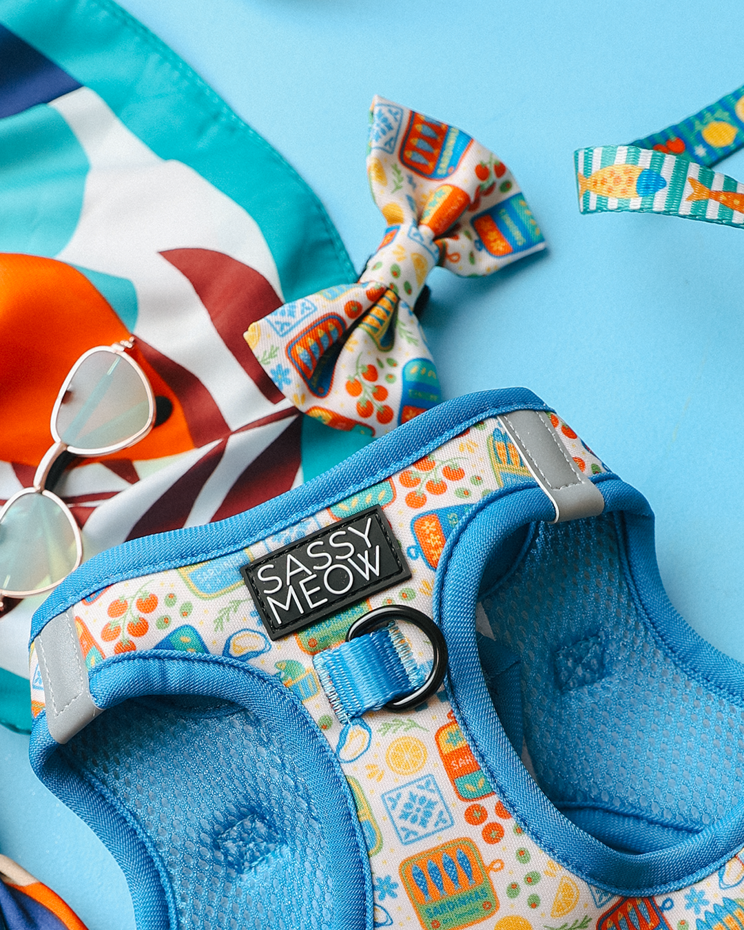 Colorful pet accessories on a blue surface, featuring the Sassy Woof Portofino Cat Step-In Harness, a matching bow tie, vibrant scarf, and stylish sunglasses.