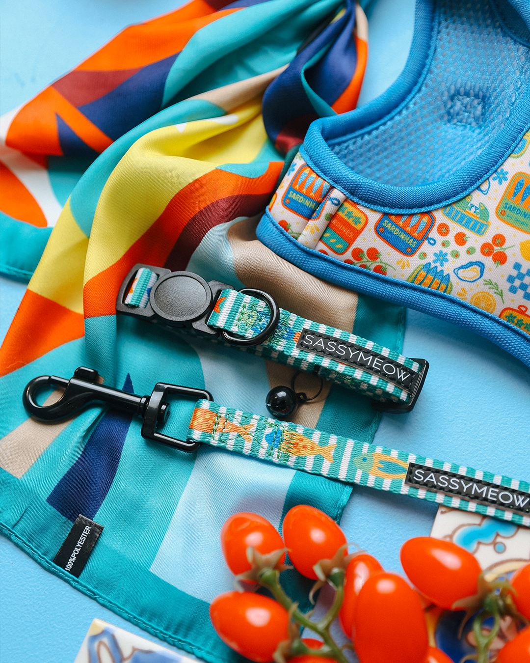 Colorful pet accessories from the Portofino Cat Collection—featuring the Sassy Woof Cat Leash and two SASSYMEOW collars—are arranged on vibrant patterned fabric, with a few red cherry tomatoes in the corner.