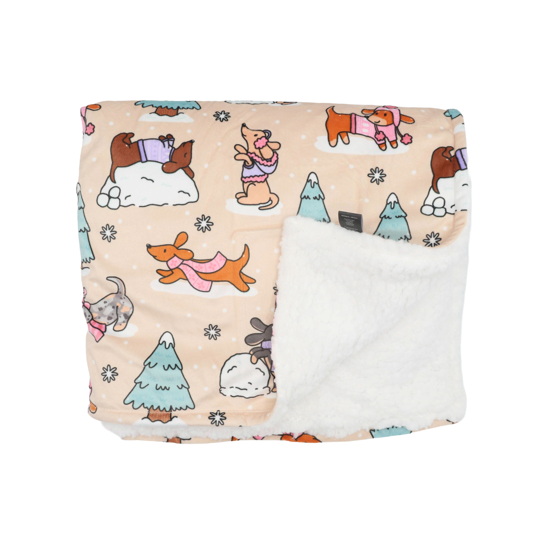 The Sassy Woof Pet Blanket - Weenie Wonderland is a beige sherpa blanket adorned with cartoon dogs in winter scarves, snowy trees, and a soft white fluffy lining—perfect as a cozy winter dog blanket—folded neatly on a white background.