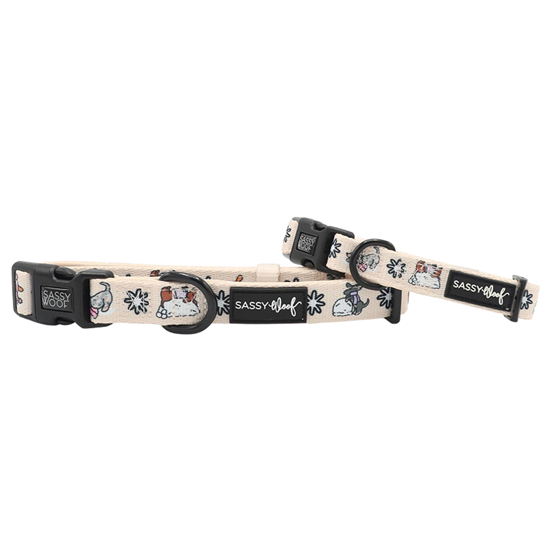 Two beige Sassy Woof “Weenie Wonderland” dog collars with black buckles and sturdy metal rings, featuring playful, colorful cartoon dogs and flowers. These festive adjustable collars are adorned with the Sassy Woof logo on black labels, making them an adorable accessory option for your pup.