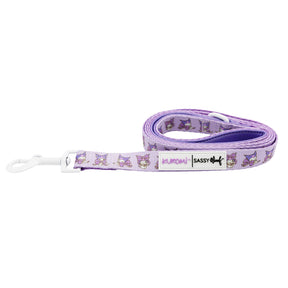 The Sassy Woof Dog Leash - Kuromi™ is purple with cartoon dog faces, white accents, a white clasp, and a "Sassy Woof" label near the handle. Pair it with a stylish harness for a playful walk. The leash comes neatly coiled.